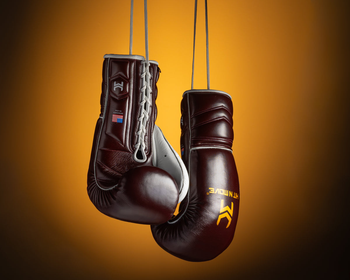 Burgundy and white HIT'N'MOVE VECTOR boxing gloves with gold logos hanging by their laces against an orange background