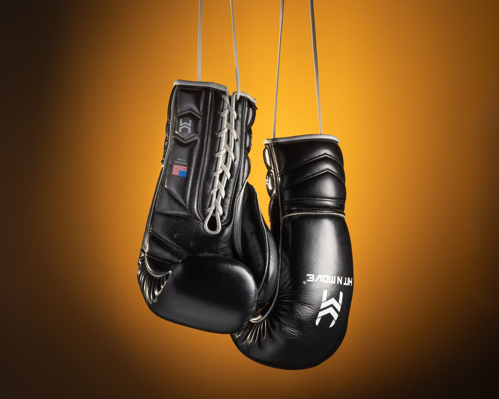 Black HIT'N'MOVE VECTOR boxing gloves with white logos hanging by white laces against an orange background