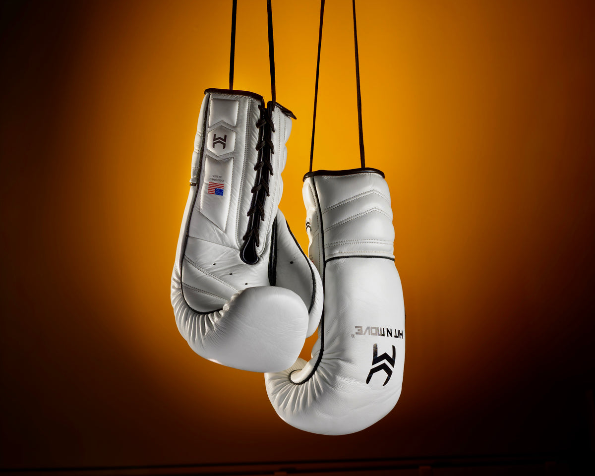 White HIT'N'MOVE VECTOR boxing gloves with black logos hanging by black laces against an orange background